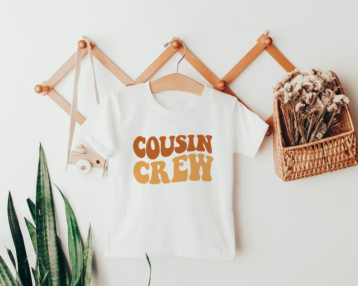 a white t-shirt with the text "COUSIN CREW" printed on it, hanging on a wooden hanger. Next to the t-shirt is a woven basket filled with flowers, and a small camera is also visible in the scene.