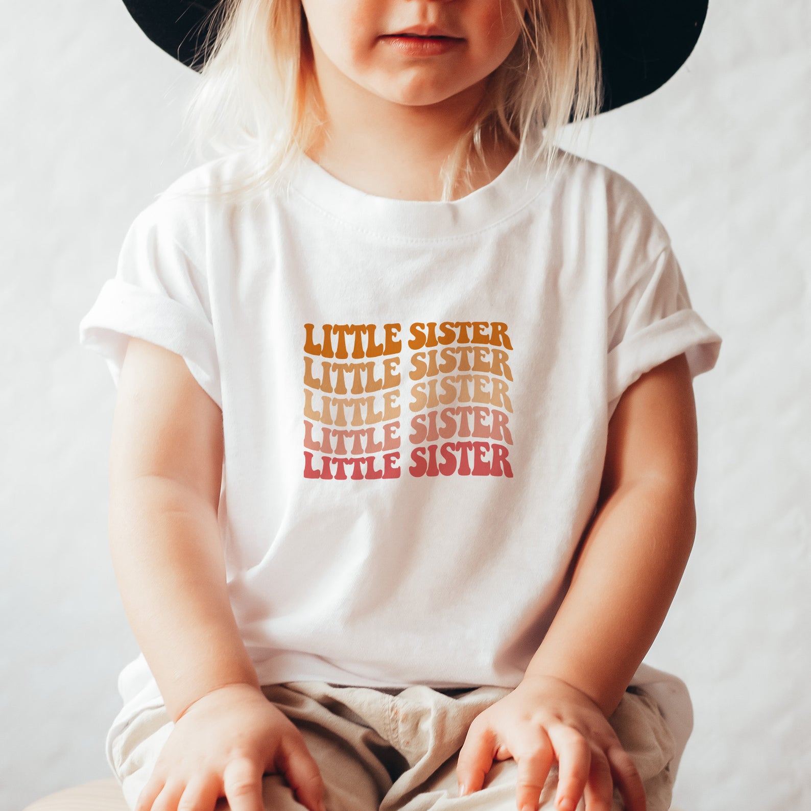 A young child wearing a white t-shirt with the text "LITTLE SISTER" printed on it.