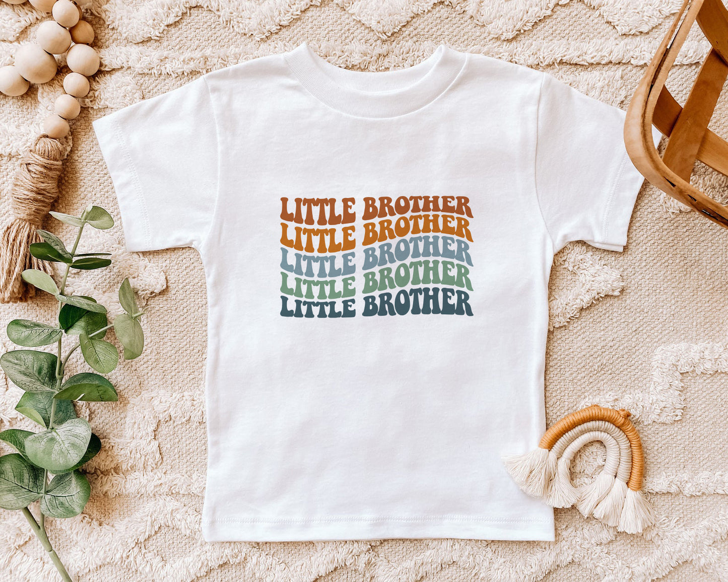 A white t-shirt with the text "LITTLE BROTHER" printed on it, surrounded by various decorative items such as a woven basket, a plant, and a rainbow-colored string.