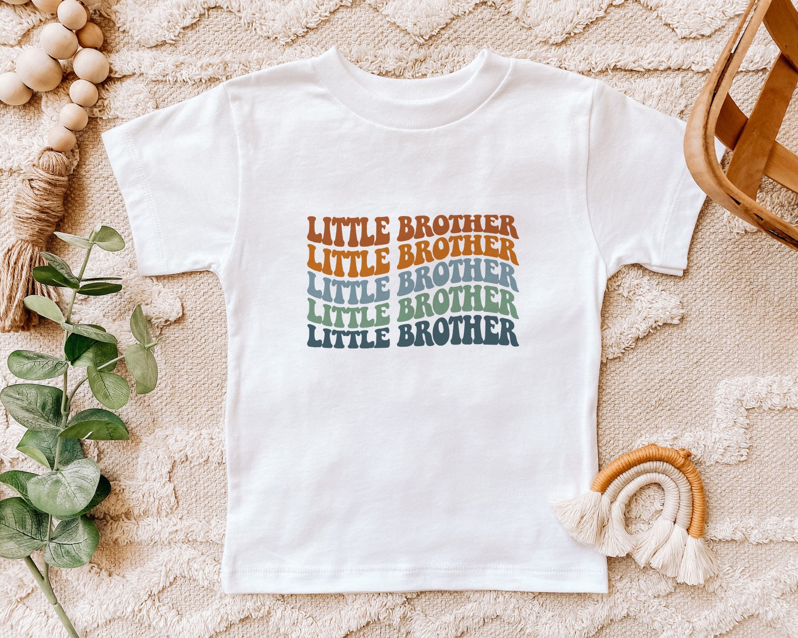 A white t-shirt with the text "LITTLE BROTHER" printed on it, surrounded by various decorative items such as a woven basket, a plant, and a rainbow-colored string.